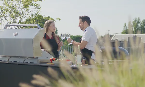 Foto einer Frau und eines Mannes, welche mit einem Glas Wein anstoßen, während Sie an der CB Outdoor Kitchen kochen.